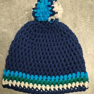 Puede incluir: Un gorro de punto azul marino con un pompón blanco y azul en la parte superior. El gorro tiene una banda de rayas verdes, azules y blancas alrededor de la parte inferior.