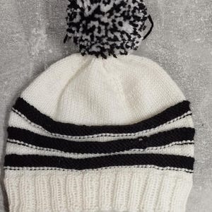 Puede incluir: Gorro de punto blanco con un pompón blanco y negro en la parte superior. El gorro tiene tres rayas negras alrededor de la parte inferior.