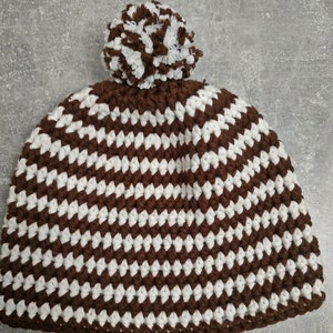 Puede incluir: Un gorro de punto de rayas marrones y blancas con un pompón blanco en la parte superior.