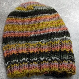 Puede incluir: Un gorro de punto a rayas en tonos de marrón, gris, amarillo, rosa y naranja. El gorro está hecho con un hilo grueso y tiene una textura acanalada.