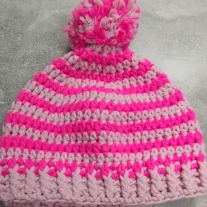 Puede incluir: Un gorro de ganchillo hecho a mano, a rayas rosas y rosa claro, rematado con un pompón esponjoso. El gorro está hecho de hilo suave y tiene un borde acanalado. El pompón es una mezcla de tonos rosas.