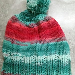Puede incluir: Un gorro de punto con un pompón en la parte superior. El gorro está a rayas en tonos de verde, rosa y rojo.