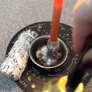 Puede incluir: Una vela naranja encendida en un pequeño frasco de vidrio, colocada en un cuenco plateado sobre una estera circular negra. Un manojo de salvia blanca y una campana dorada sugieren un tema espiritual o ritual.