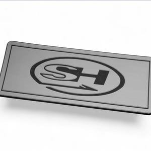 May include: A rectangular, silver metal plate with a black border. The plate features a black oval design in the centre, containing the letters "SH" and an arrow. The design is slightly raised, creating a subtle 3D effect.