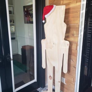 Puede incluir: Un recorte de madera de una persona que lleva un gorro de Papá Noel rojo. El recorte está de pie sobre una base de madera y se apoya en una pared con paneles de madera.