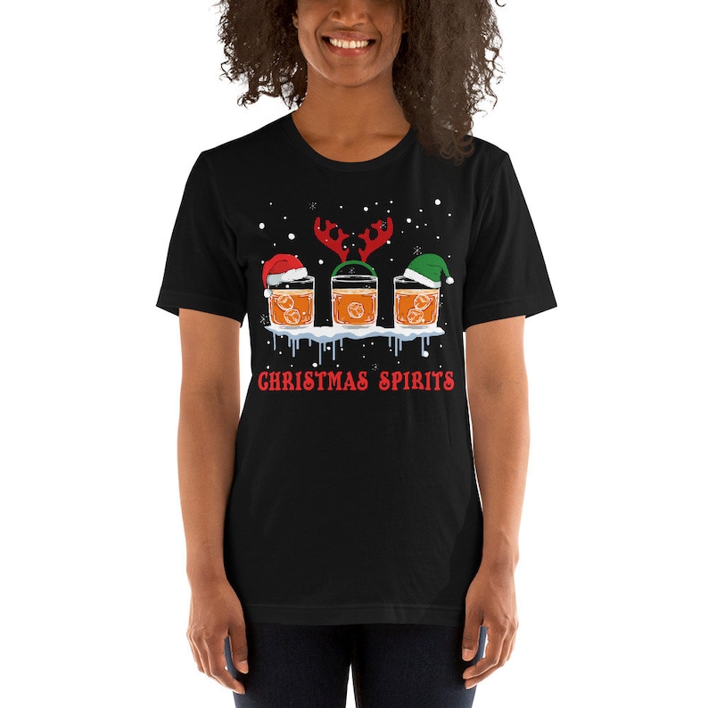 May include: Black t-shirt with a Christmas-themed design featuring three glasses of whiskey with Santa hats and reindeer antlers. The text "Christmas Spirits" is printed below the glasses.