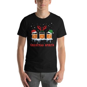 May include: A black t-shirt with a Christmas-themed design featuring three glasses of whiskey with Santa hats and reindeer antlers. The text "Christmas Spirits" is printed below the glasses.