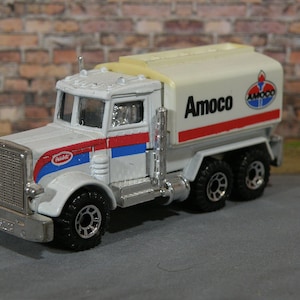 Puede incluir: Un camión de juguete blanco con un esquema de pintura rojo, blanco y azul. El camión tiene un gran tanque de combustible con la palabra "Amoco" impresa en él. El camión es un modelo de juguete de un Peterbilt.