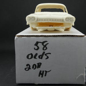 Könnte beinhalten: Ein cremefarbenes Modellauto, vermutlich ein Oldsmobile aus der Zeit, steht auf einem Karton. Das Vorderteil des Autos ist sichtbar, mit Scheinwerfern und dem Wort "OLDSMOBILE" auf der Vorderseite. Die Box ist mit den Zahlen "58 olds 2dr HT" beschriftet.