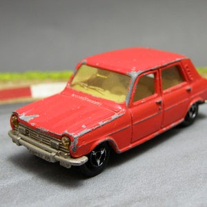 Può includere: Modello di auto giocattolo rosso di una Simca 1100, un'auto compatta francese, con una finitura della vernice leggermente usurata. L'auto è su una superficie grigia.