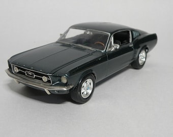 以下が含まれることがあります： 濃い緑色の1967年型フォード・マスタングのモデルカー。車は黒い内装とクロムのアクセントが施されています。車は白い表面の上に置かれています。