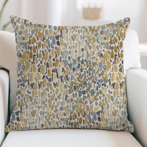 May include: Square throw pillow with a watercolor pattern. The design features an abstract arrangement of small, elongated shapes in shades of gold, blue, and gray on a white background. The pillow is placed on a white armchair.