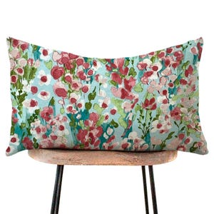 May include: Rectangular decorative pillow with a floral pattern. The pillow features a light blue background with a watercolor design of red, pink, and green flowers. The pillow is resting on a small wooden stool with black metal legs.