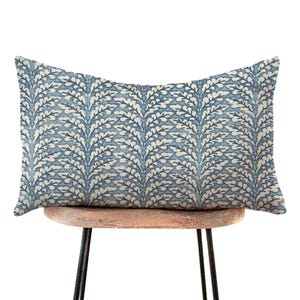 May include: A rectangular decorative pillow with a blue and white leaf pattern. The pillow is resting on a small, round wooden stool with black metal legs. The pillow's design features a repeating pattern of stylized leaves.