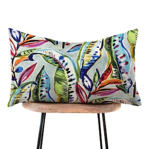 May include: Rectangular decorative pillow with a colorful tropical leaf pattern. The pillow features a light blue background with vibrant green, blue, orange, and purple leaves. The pillow is placed on a small wooden stool with black metal legs.