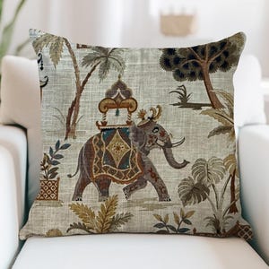 May include: Decorative square pillow featuring an elephant with a decorative saddle, walking through a landscape with palm trees and other foliage. The pillow has a neutral background with brown, blue, and gold accents. The pillow is on a white couch.