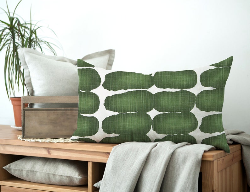 May include: A green and white rectangular throw pillow with an abstract pattern. The pillow is sitting on a wooden surface with a blanket draped over it.
