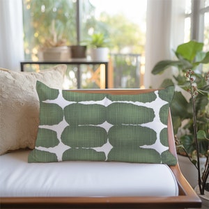 May include: A green and white patterned throw pillow with a geometric design. The pillow is sitting on a white sofa with a wooden frame.