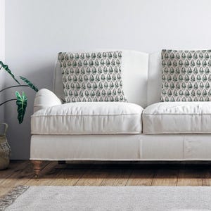 Puede incluir: Un sof&aacute; blanco con dos cojines con un patr&oacute;n geom&eacute;trico verde y blanco. El sof&aacute; est&aacute; en un sal&oacute;n con una planta verde en una cesta de mimbre.