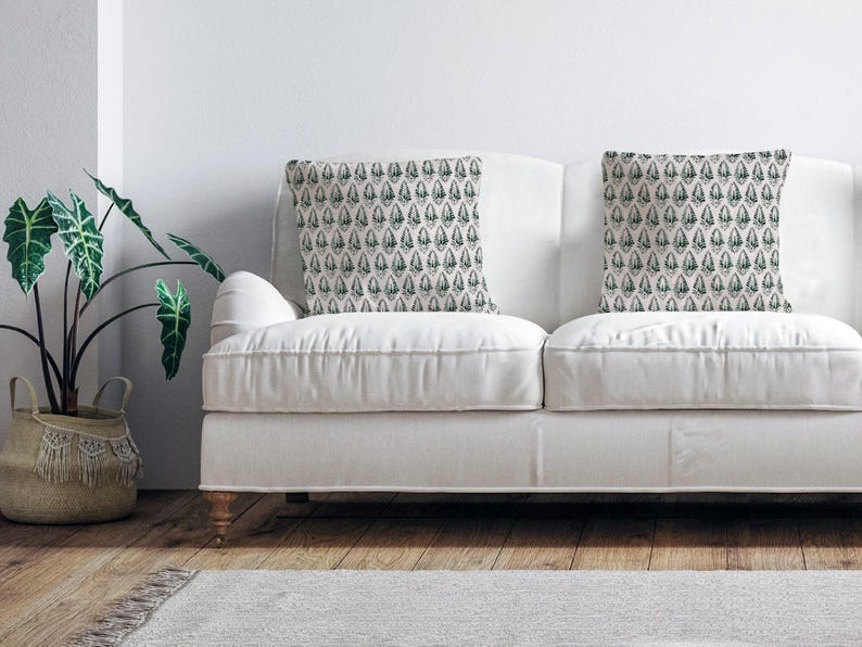 Puede incluir: Un sof&aacute; blanco con dos cojines con un patr&oacute;n geom&eacute;trico verde y blanco. El sof&aacute; est&aacute; en un sal&oacute;n con una planta verde en una cesta de mimbre.