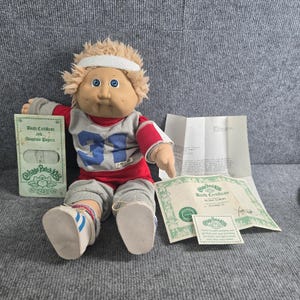 Puede incluir: Una muñeca Cabbage Patch Kids con cabello rubio, una cinta blanca y una camiseta roja y gris con el número 31. La muñeca viste pantalones grises y zapatillas blancas con rayas azules, acompañada de un certificado de nacimiento y papeles.