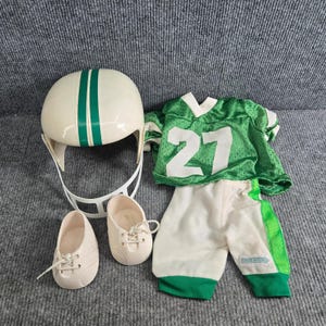 May include: A doll-sized football uniform set including a cream-colored helmet with two vertical green stripes and a white face mask, a green mesh jersey with the white number 27, white pants with green side stripes, and two cream-colored lace-up shoes.