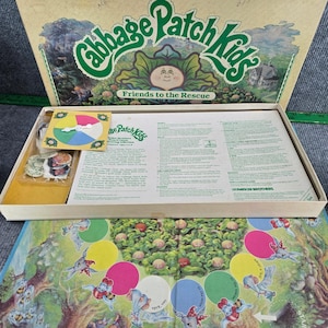 Può includere: Un set di gioco da tavolo Cabbage Patch Kids Friends to the Rescue con un coperchio della scatola con testo verde e un'illustrazione di cavolo, un tabellone con cerchi colorati, una ruota con una freccia bianca e un foglio di istruzioni.