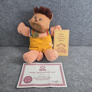 Può includere: Un peluche a forma di gatto Cabbage Patch Kids Koosas con capelli castani, arti a righe arancioni e bianche e salopette in velluto a coste giallo. Indossa un collare marrone con una medaglietta in metallo. Include un certificato di identificazione.