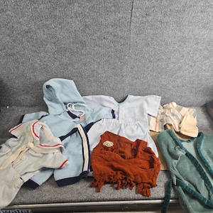May include: A collection of doll clothing including a light blue hooded sweatshirt with dark blue trim, a light blue short-sleeved shirt, a white skirt, a rust-colored fringed top, a striped shirt, and a green corduroy vest with matching pants.