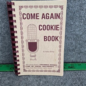 Könnte beinhalten: Ein beigefarbenes spiralgebundenes Kochbuch mit dem Titel COME AGAIN COOKIE BOOK von Evelyn Birkby, das eine dunkelmarone Mikrofonillustration mit dem Text KMA zeigt. Der Einband hat einen dekorativen Rand und liegt neben einem grünen Lineal.