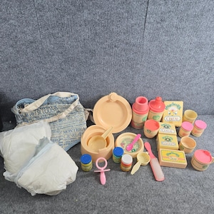 May include: A collection of plastic toy baby care accessories including a blue and white patterned fabric bag, white diapers, a peach-colored plate, bowls, bottles, a pink pacifier, a hairbrush, and small boxes labeled Cereal, Cookies, and Soap.