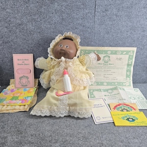 Op de afbeelding: Een Cabbage Patch Kids pop met een donkere huidskleur, gekleed in een gele jurk met kanten rand en een bijpassende muts. De pop houdt een klein wit flesje met een roze dop vast. Inbegrepen zijn diverse papieren documenten, waaronder een officieel adoptiebewijs en geboortecertificaat, samen met een kleine geïllustreerde kaart en een gele informatiebrochure.