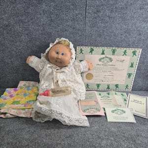 Puede incluir: Una muñeca Cabbage Patch Kids con ojos azules y cabello de lana, vestida con un gorro y vestido blanco con encaje. La muñeca sostiene un biberón de plástico transparente con tapa rosa. Incluye certificado de adopción y tarjetas oficiales.