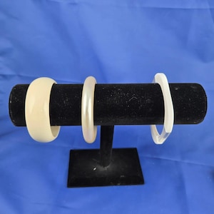 May include: Three bangles displayed on a black stand against a blue backdrop. The bangles are in shades of cream, pearl, and clear. The cream bangle is thick and rounded, the pearl bangle is thin, and the clear bangle has a geometric shape.