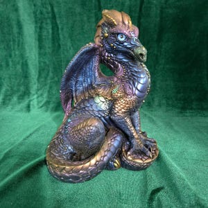 Peut inclure: Figurine de dragon fantastique détaillée dans les tons de violet, bleu et or. Le dragon est assis, les ailes déployées, mettant en valeur les détails complexes des écailles et de la texture. Le dragon a les yeux bleus et se détache sur un fond vert.