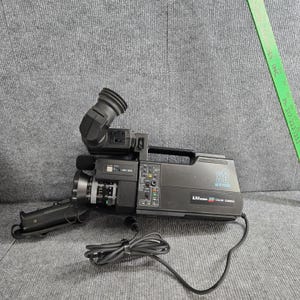 May include: A black LXI Series X8 Auto Focus color video camera with a top handle, a side-mounted viewfinder, and a lens. A green ruler is visible in the upper right corner, showing measurements in inches. A black power cord is attached to the base.