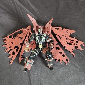 Peut inclure: Une figurine d'action noire et rouge de Spawn, un super-héros avec un crâne sur la poitrine, portant une cape au design déchiré, rouge et noir.