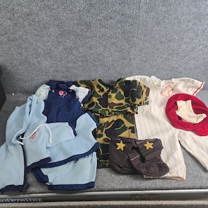 Puede incluir: Una colección de ropa para muñecos que incluye una chaqueta con capucha y pantalones cortos azul claro, un conjunto de peto azul marino con bordado de globos, una camisa y pantalones de camuflaje, un uniforme de béisbol a rayas blancas, una gorra roja y calcetines oscuros con estrellas.