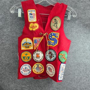 May include: A red felt vest adorned with numerous colorful patches. The patches feature text and graphics related to scouting, including the words "Recruiter," "Trailsend," and "Great Rivers Council." The vest is suspended from a clear hanger.