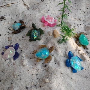 Op de afbeelding: Een verzameling van zeven kleine schildpadfiguren van hars op een zanderig oppervlak. De schildpadden zijn in verschillende kleuren, waaronder blauw, groen, paars en bruin, en hebben iriserende glitteraccenten. Eén schildpad is roze en heeft de vorm van een zeepaardje.