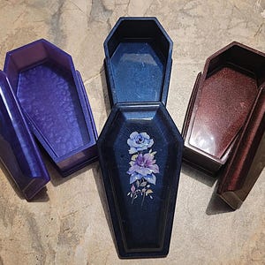 May include: Three coffin-shaped boxes made of resin. One is purple with a textured interior, one is dark blue with a floral rose decal on the lid, and one is a deep metallic red. Each box features a removable lid and a hollow interior compartment.
