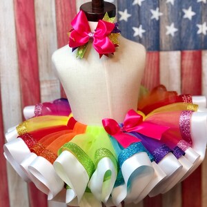May include: Rainbow tulle tutu skirt with white satin ribbon trim and a pink bow. The skirt has a layer of rainbow tulle with glitter accents.