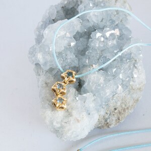 Puede incluir: Un delicado collar de cordón azul con un colgante de oro con tres abalorios en forma de estrella con piedras preciosas azules.
