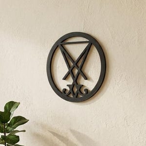 May include: A circular black wall hanging featuring the Sigil of Lucifer, a geometric design with a central cross and V-shaped lines enclosed in a ring. The piece is mounted on a light-colored wall above a wooden console table with a plant and books.