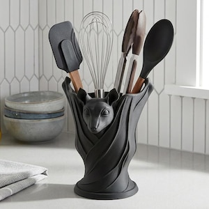 May include: A matte black kitchen utensil holder sculpted in the shape of a bat with wings wrapped around the base. The holder contains a metal whisk, a black silicone spatula, a pair of brown-handled tongs, and a black silicone serving spoon.