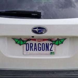May include: A white Ohio license plate with the text DRAGON2 in blue, framed by two black and green dragon wing attachments mounted on the top corners of the plate, installed on the rear hatch of a white Subaru Outback vehicle.