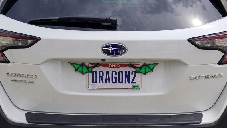 May include: A white Ohio license plate with the text DRAGON2 in blue, framed by two black and green dragon wing attachments mounted on the top corners of the plate, installed on the rear hatch of a white Subaru Outback vehicle.