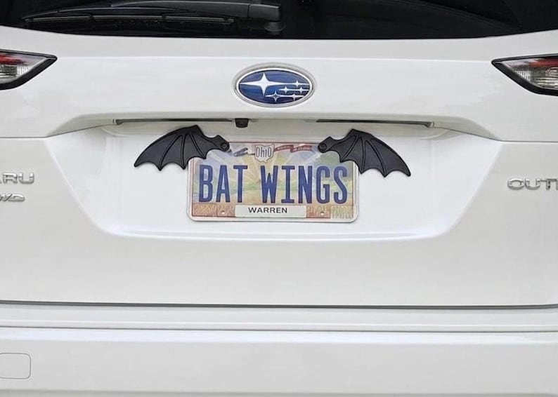 May include: Black bat wing car accessories mounted above a license plate that reads "BAT WINGS" in blue letters. The license plate is from Warren, Ohio. The car is white.