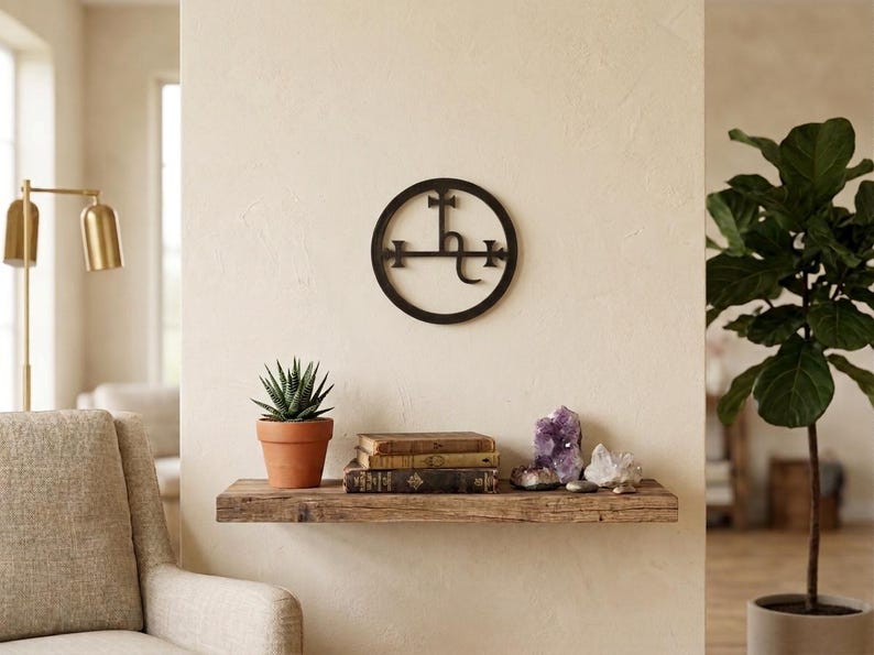 May include: A circular black metal wall art piece featuring a central alchemical symbol of Saturn, mounted on a light-colored wall above a rustic wooden floating shelf that holds a potted succulent, stacked vintage books, and raw crystal clusters.