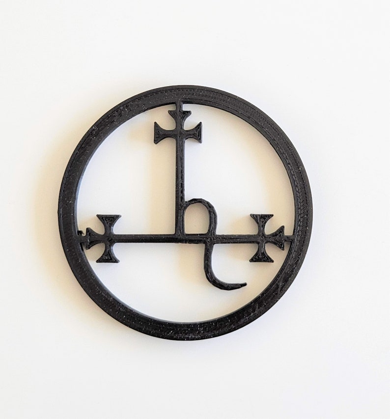 May include: A circular black wall decoration featuring an intricate symbol inside. The design consists of a central vertical line topped with a cross, intersected by a horizontal bar with crosses at each end, and a curved tail at the bottom.